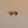 Amethyst Heart Hoops featuring purple heart-shaped stones and small clear gems, reminiscent of diamond huggies, displayed on a beige textured surface.
