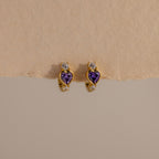 Amethyst Heart Hoops featuring purple heart-shaped stones and small clear gems, reminiscent of diamond huggies, displayed on a beige textured surface.