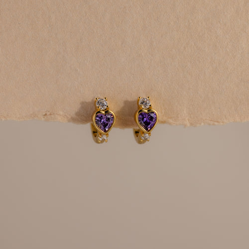 Amethyst Heart Hoops featuring purple heart-shaped stones and small clear gems, reminiscent of diamond huggies, displayed on a beige textured surface.
