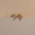 The Alexandrite Heart Hoops feature gold tones, purple heart-shaped stones, and small clear gems, capturing the charm of birthstone earrings. Their sparkle stands out beautifully against a beige background.