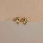 Diamond Heart Hoops featuring heart-shaped and round clear gemstones are elegantly displayed on a beige textured background.