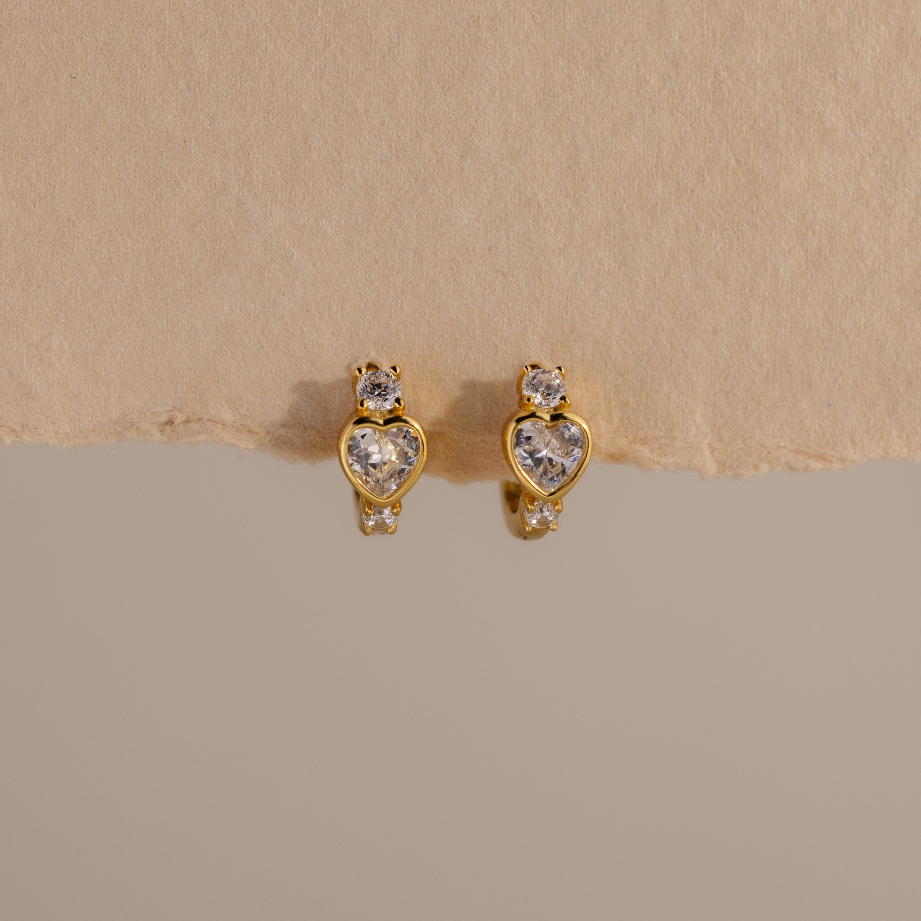 Diamond Heart Hoops featuring heart-shaped and round clear gemstones are elegantly displayed on a beige textured background.