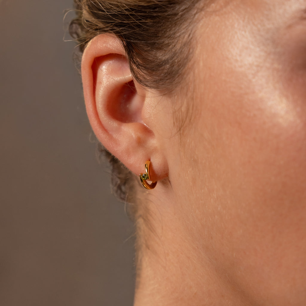 Close-up of a womans ear wearing Floating Emerald Huggies—gold hoops with a subtle green gem, perfect for fans of personalized jewelry.