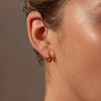 Close-up of a womans ear wearing Floating Sapphire Huggies—small gold hoop earrings accented with a blue gemstone for a personalized jewelry touch.