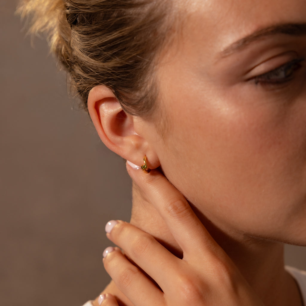 Close-up of a woman touching her neck, showcasing Floating Peridot Huggies—delicate gold earrings that add a subtle, elegant touch to any look.