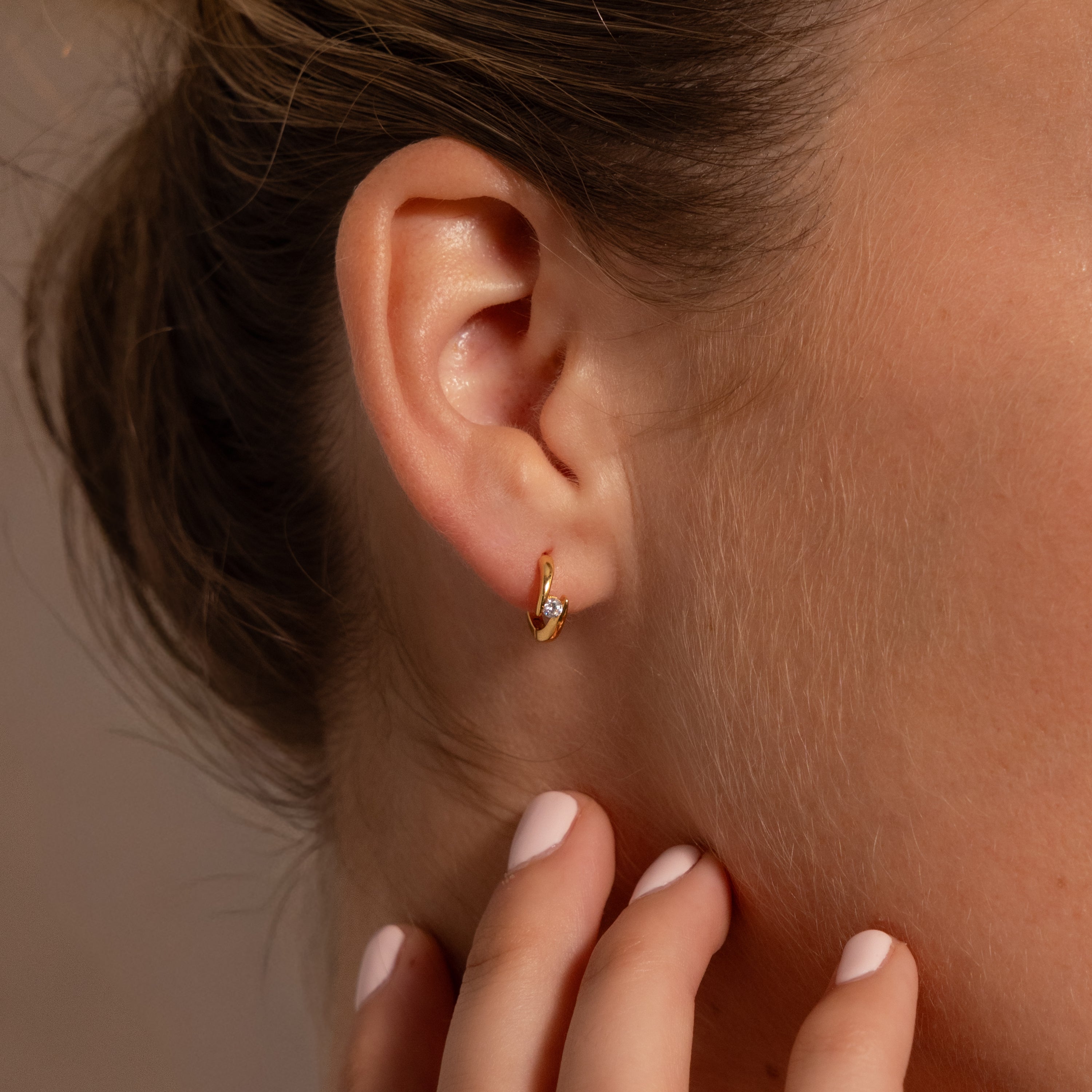 Close-up of a womans ear wearing Floating Topaz Huggies, her hand touching her neck and showcasing elegant personalized gold hoop earrings.