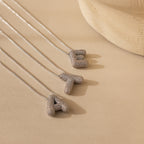 Three Pave Puff Letter Necklaces in White Gold—featuring textured pave A, T, and E pendants—rest on a beige surface.