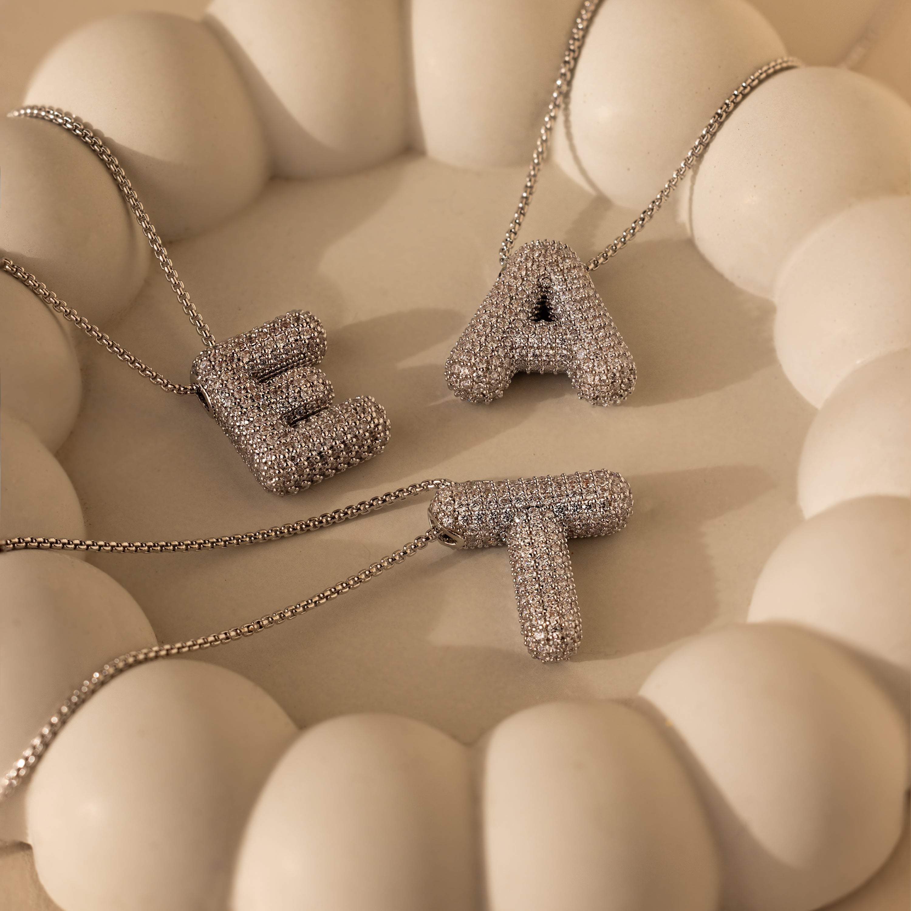 Three Pave Puff Letter Necklaces with sparkling E, A, and T pendants are displayed on a cream, rounded tray, adding elegance to your collection.