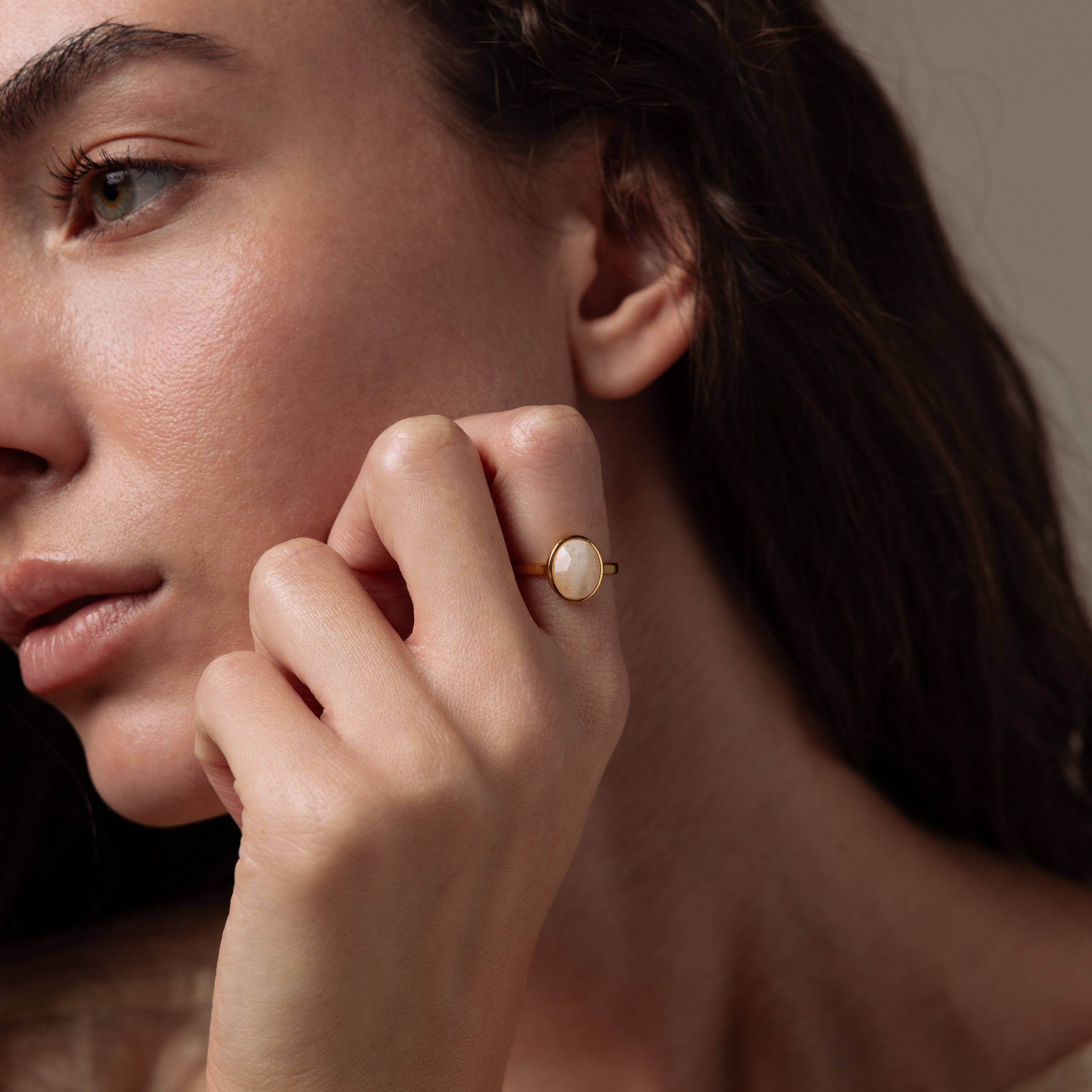 Woman with long brown hair wears the Moonstone Signet Ring, a minimalist gold band with a round white moonstone, holding her hand near her face—an elegant everyday jewelry piece.