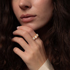 Woman with wavy brown hair wearing the Cosmic Moonstone Ring, a gold statement piece with white stones—her hand near her face adds a chic boho touch.