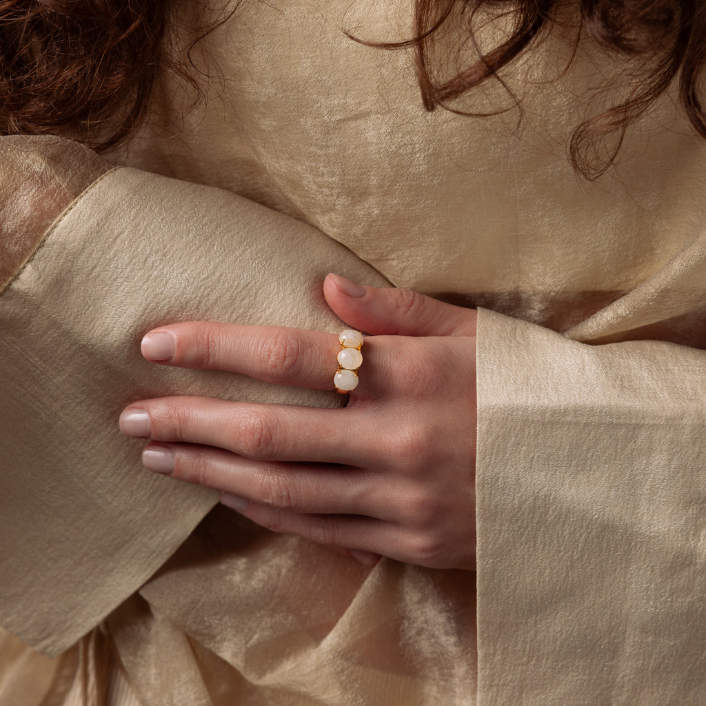 A person in a beige, sheer garment displays a hand adorned with the Cosmic Moonstone Ring, a gold statement piece featuring three white stones that embody boho elegance.