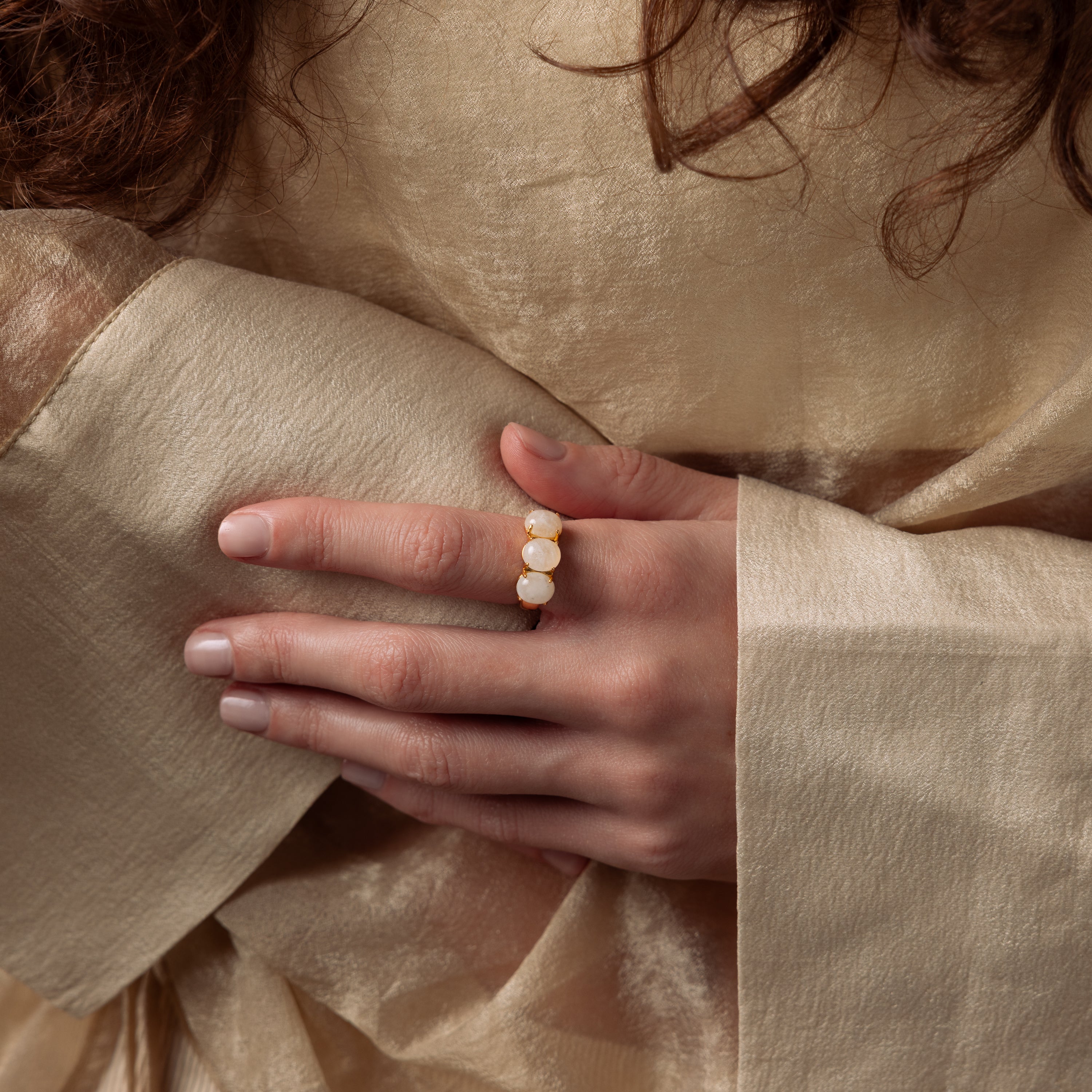 A person in a beige, sheer garment displays a hand adorned with the Cosmic Moonstone Ring, a gold statement piece featuring three white stones that embody boho elegance.