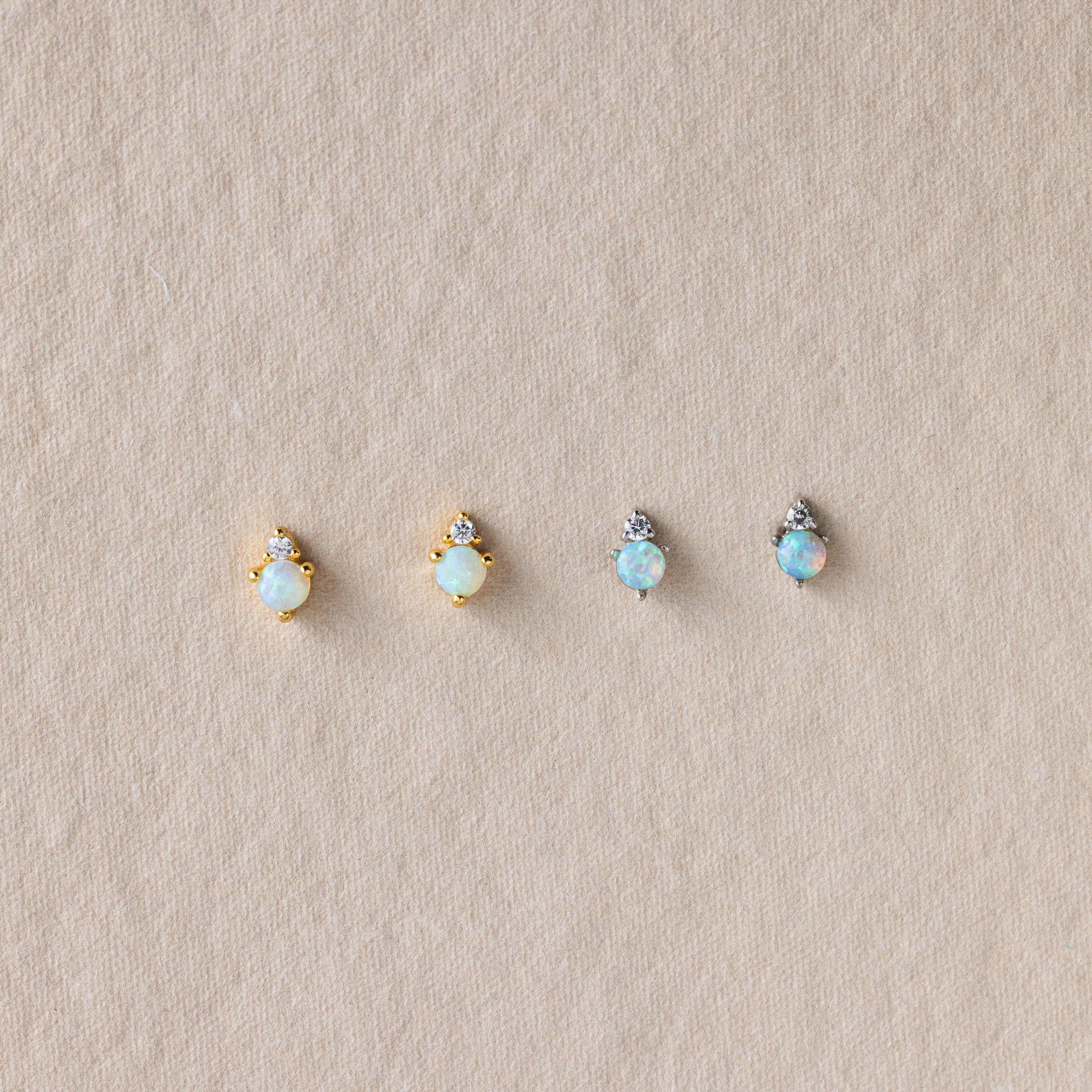 Four dainty Raindrop Emerald Studs with gold and silver settings are displayed on a textured beige surface.