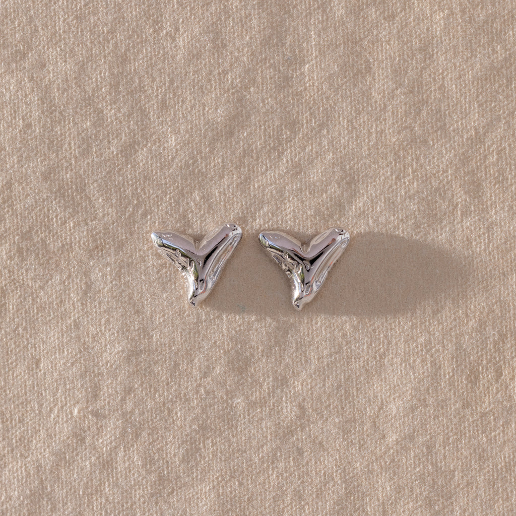 The Shark Tooth Flatback Studs in Silver rest on a textured beige surface—a stylish beach accessory to elevate your look.