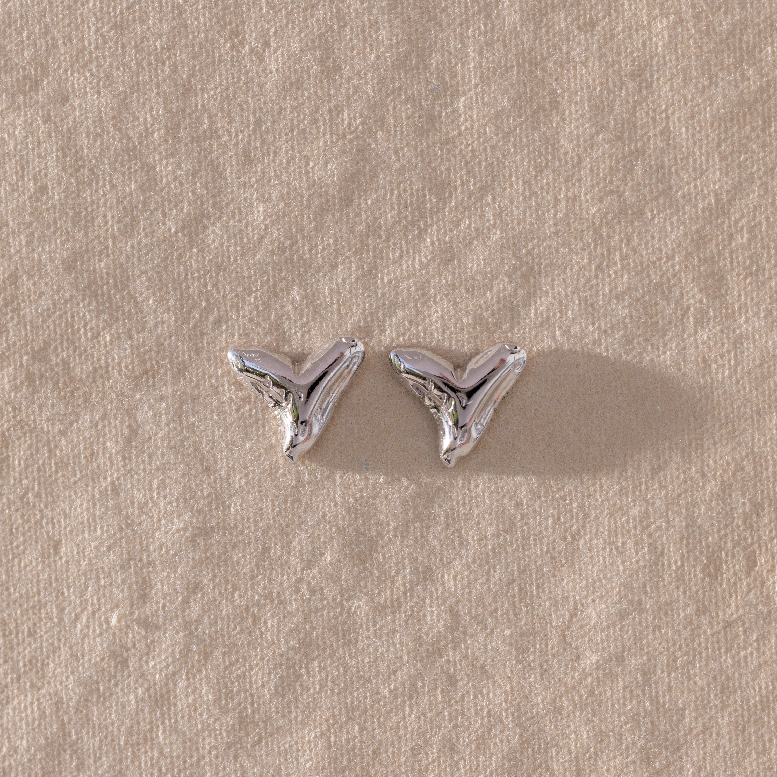 The Shark Tooth Flatback Studs in Silver rest on a textured beige surface—a stylish beach accessory to elevate your look.
