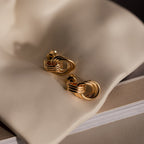 The Micro Triple Knot Earrings in 18K Gold embody quiet luxury as they rest on soft beige fabric, with a closed book beneath them.