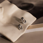 Micro Triple Knot Earrings in Silver shown on a wavy ceramic tray, with a blurred book in the background.