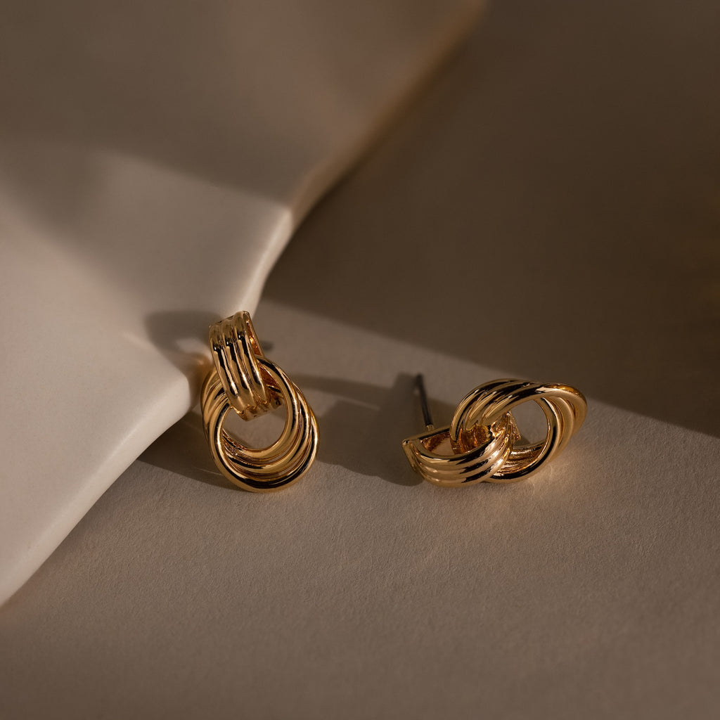 Micro Triple Knot Earrings in 18K Gold rest on a neutral surface, softly illuminated to highlight their effortless quiet luxury.