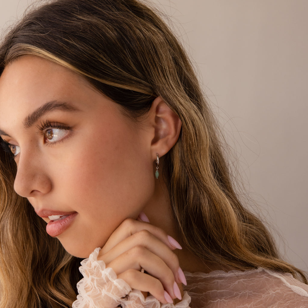 Woman with long brown hair, natural makeup, wearing a sheer ruffled top and Jade Marquise Huggies in Silver earrings, looking left.