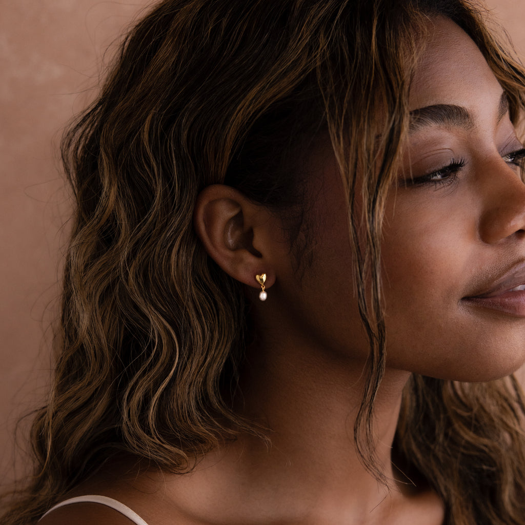 Woman with wavy hair smiles softly, wearing elegant Heart Pearl Drop Earrings in 18K Gold against a neutral background.
