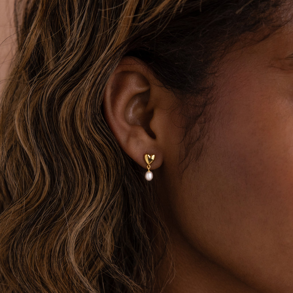 Close-up of the Heart Pearl Drop Earrings in 18K Gold, showcasing a gold heart-shaped stud with a small pearl, worn on a womans ear with wavy hair.
