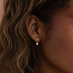 Close-up of the Heart Pearl Drop Earrings in 18K Gold, showcasing a gold heart-shaped stud with a small pearl, worn on a womans ear with wavy hair.