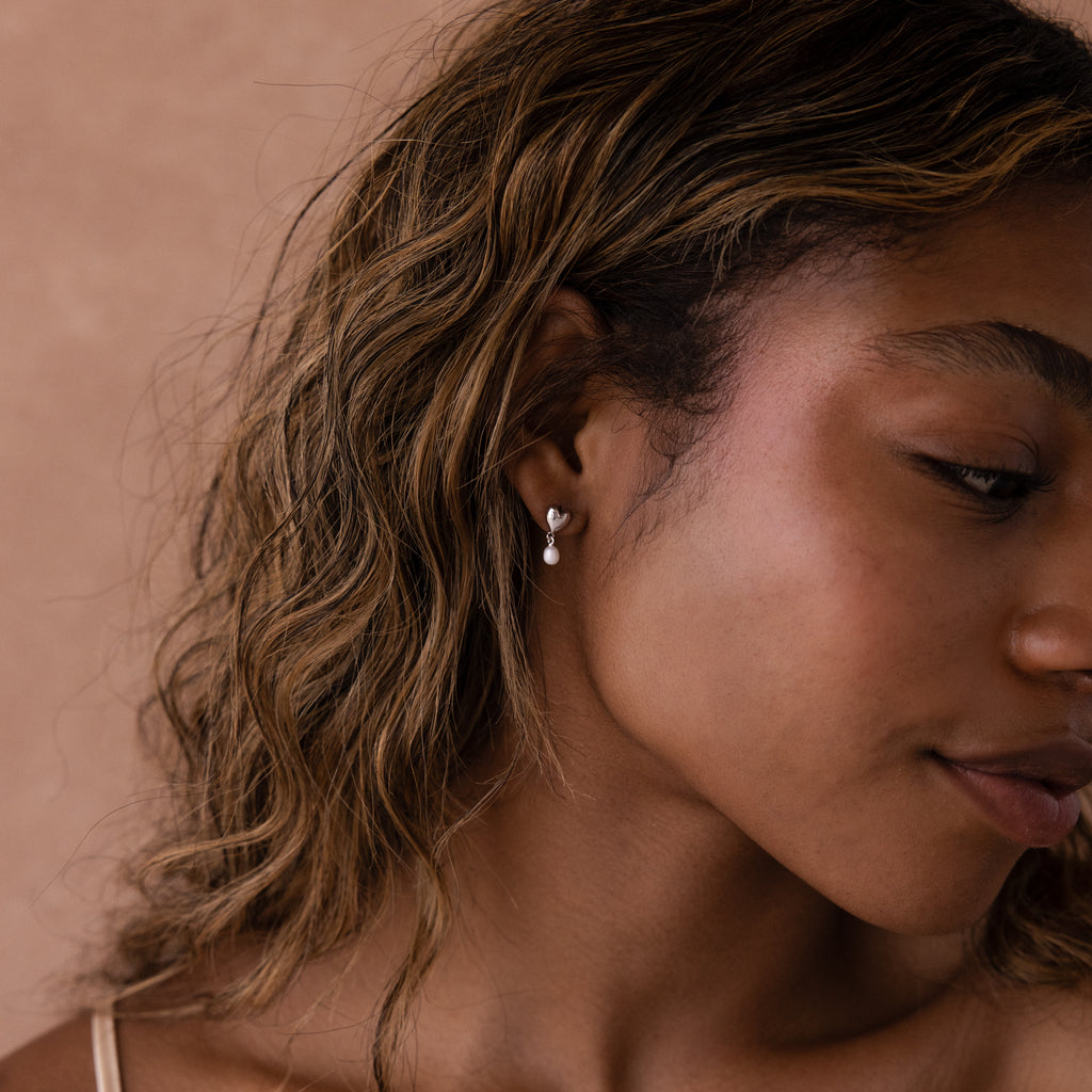 Close-up of a woman with wavy hair wearing Heart Pearl Drop Earrings in Silver against a neutral background.