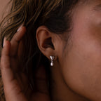 A close-up of a person wearing Heart Pearl Drop Earrings in Silver, featuring heart shapes and dangling pearls for an elegant look.
