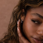 A woman with long wavy hair touches her face, highlighting the elegant Heart Pearl Drop Earrings in Silver, featuring a delicate heart-shaped pearl design.