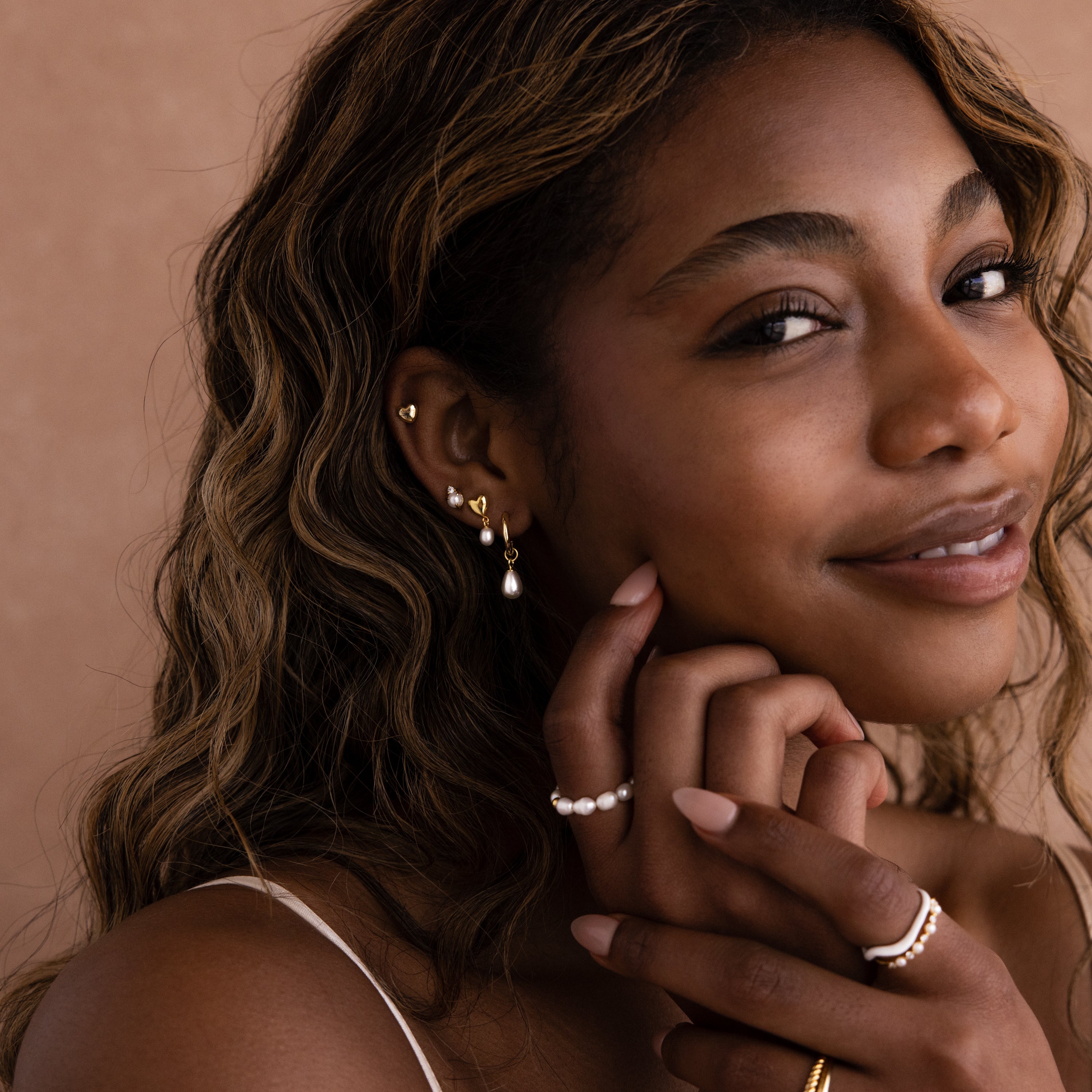 Woman with wavy hair smiles, wearing gold earrings, Heart Pearl Drop Earrings in 18K Gold, a pearl ring, and several gold rings against a beige background.