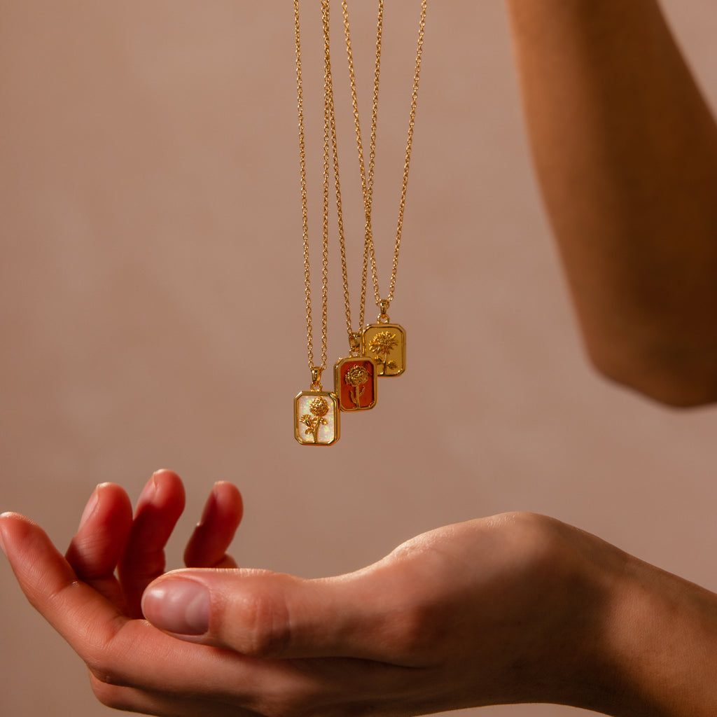 The Topaz Birth Flower Charm Necklace is displayed above an outstretched hand against a neutral, minimalist background—perfect for showcasing elegant jewelry.