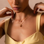 A woman in a yellow dress elegantly adjusts two gold Birth Flower Charm Necklaces, each featuring a small pendant.