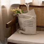 The CM Tote Bag in beige, featuring a minimalist design, is shown with tulips placed on a wooden chair as sunlight filters through sheer curtains in the background.