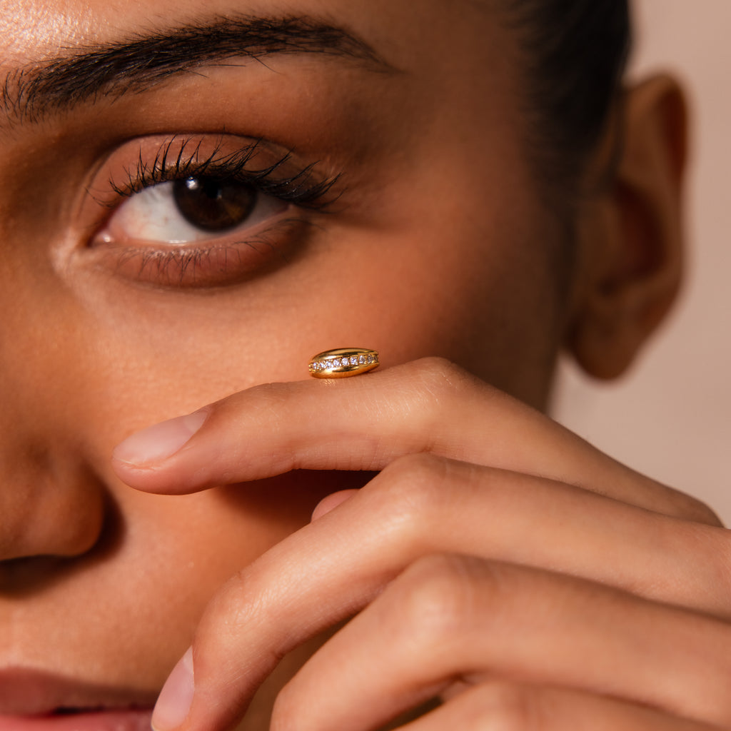 A woman holds the Pave Dome Charm in 18K Gold near her face, its sparkling design catching the light and highlighting her eye with graceful elegance.