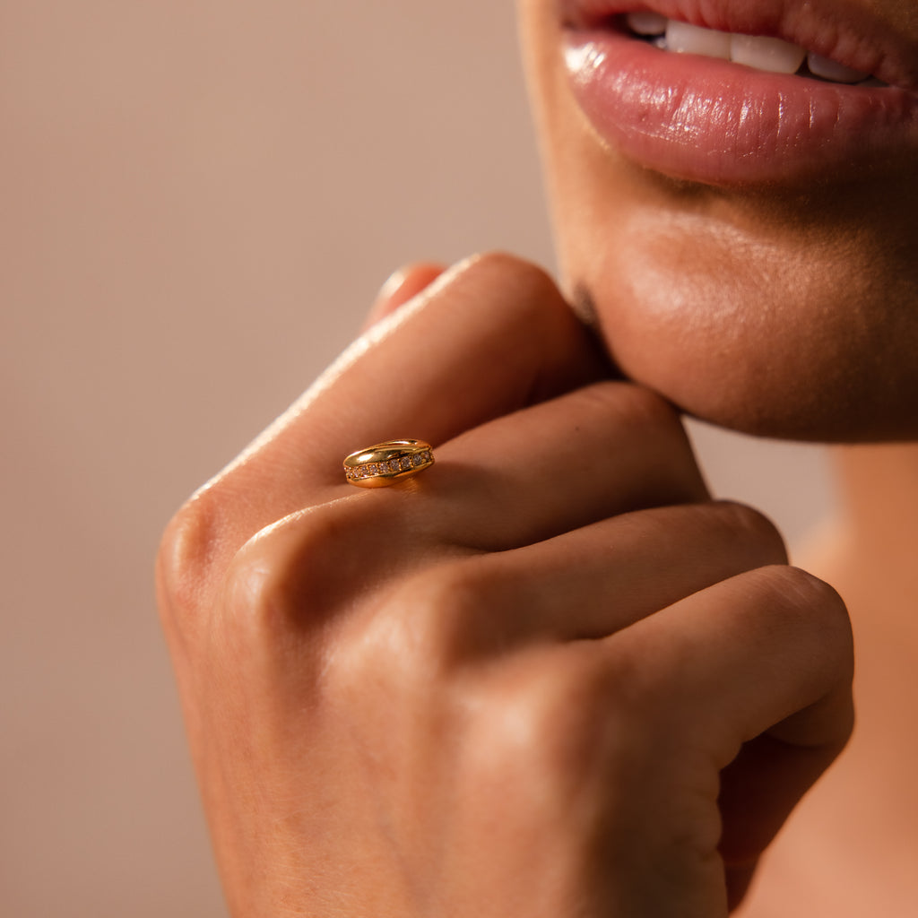 A close-up of a woman's hand near her lips, wearing a gold ring with sparkling diamonds—a stunning piece to enhance any charm stack or perfectly paired with the Pave Dome Charm in 18K Gold.