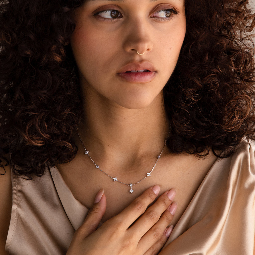 A woman with curly hair wears the Ivy Diamond Station Necklace in Silver and a beige satin top, her hand resting on her chest.