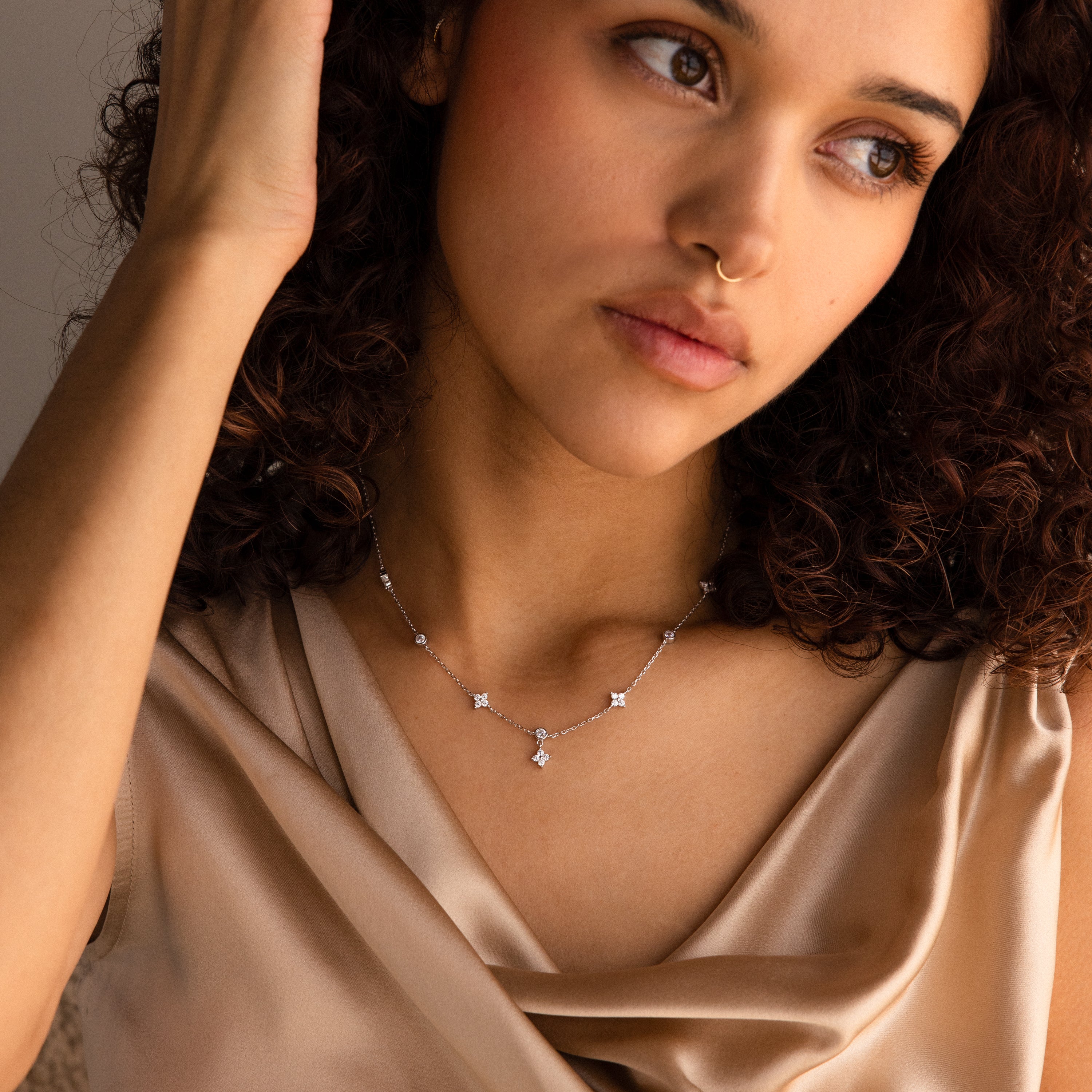 Woman with curly hair wearing a satin top, a nose ring, and the Ivy Diamond Station Necklace in Silver, looking thoughtful.