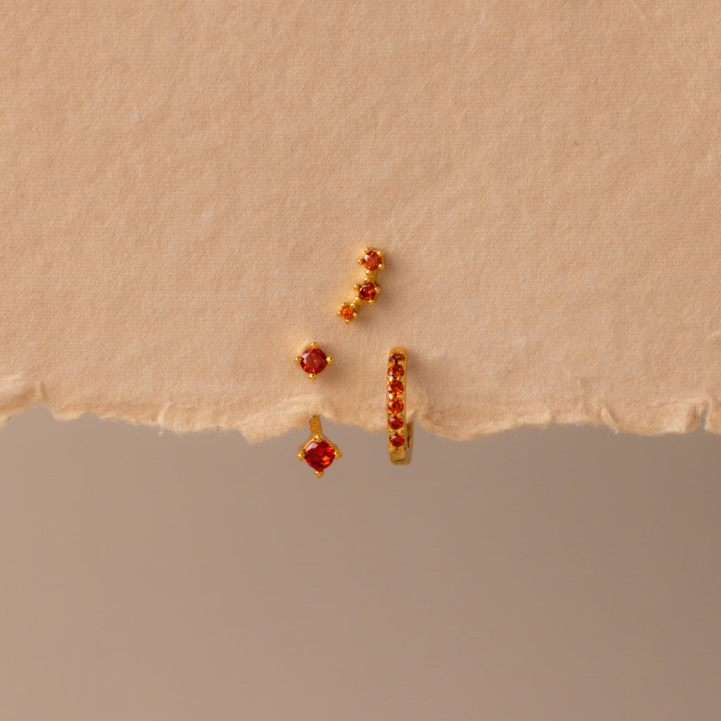 The Garnet Earring Trio Set—three gold earrings with red gemstones, including a huggie hoop—are arranged on torn beige paper for a striking display.