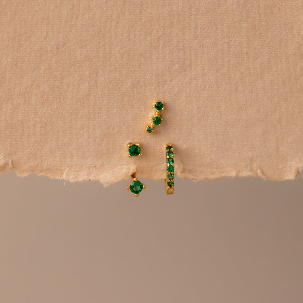 The Emerald Earring Trio Set features gold earrings and studs with green gemstones, including an elegant birthstone pair and pavé huggie hoops, displayed on a textured beige surface.