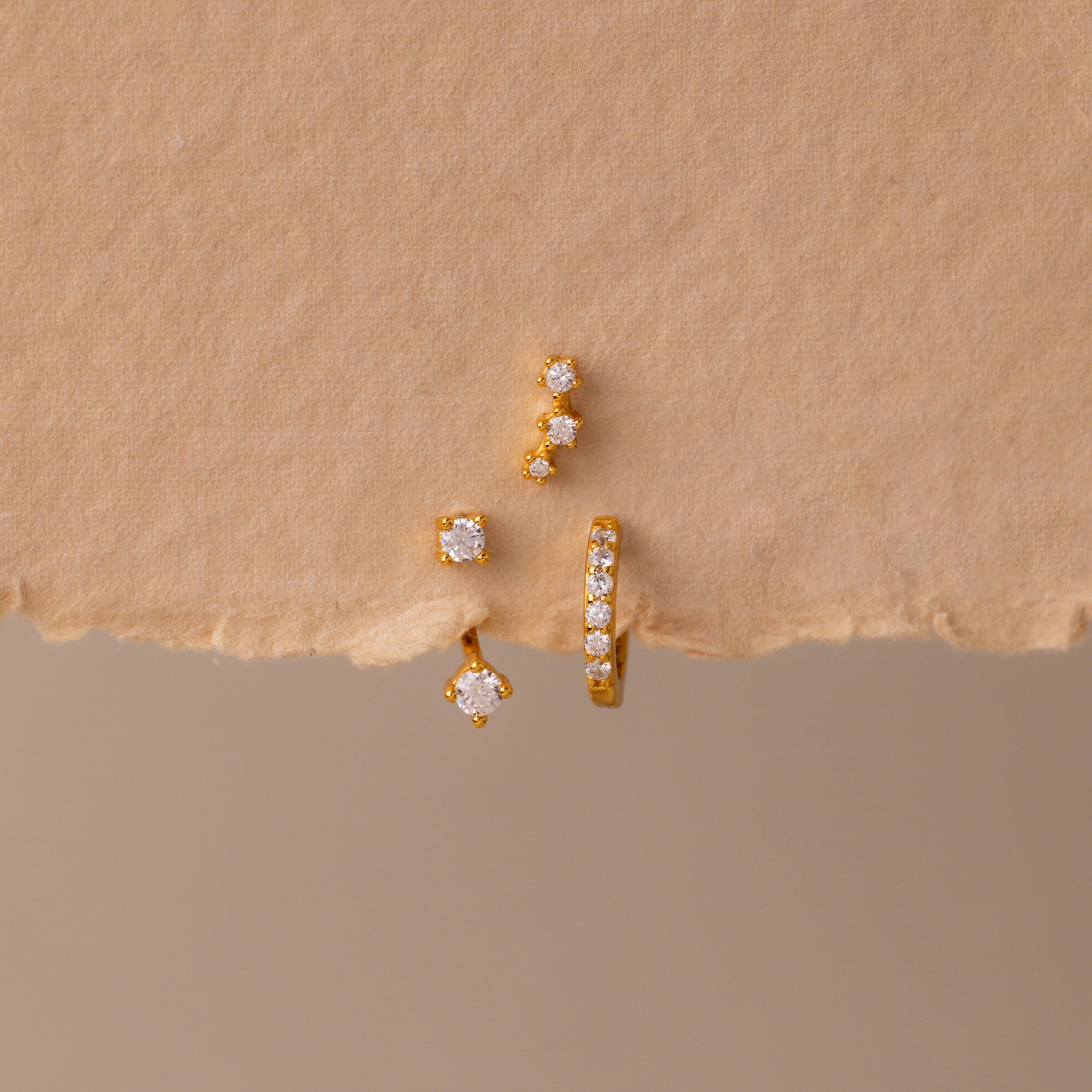 The Diamond Earring Trio Set sparkles elegantly with small white gemstones on a textured beige surface, ideal for anyone who loves versatile birthstone earrings.
