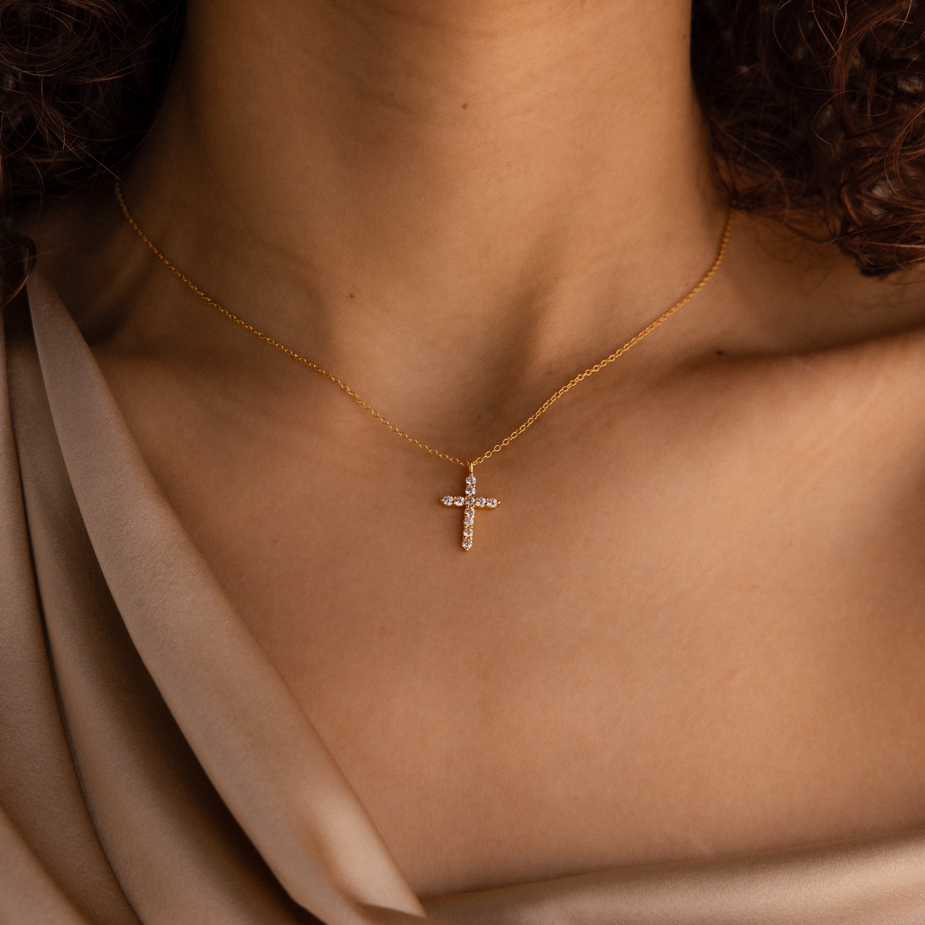A woman wearing the Pave Cross Necklace in 18K Gold, featuring a jeweled cross pendant on a gold chain, paired with a beige satin top.