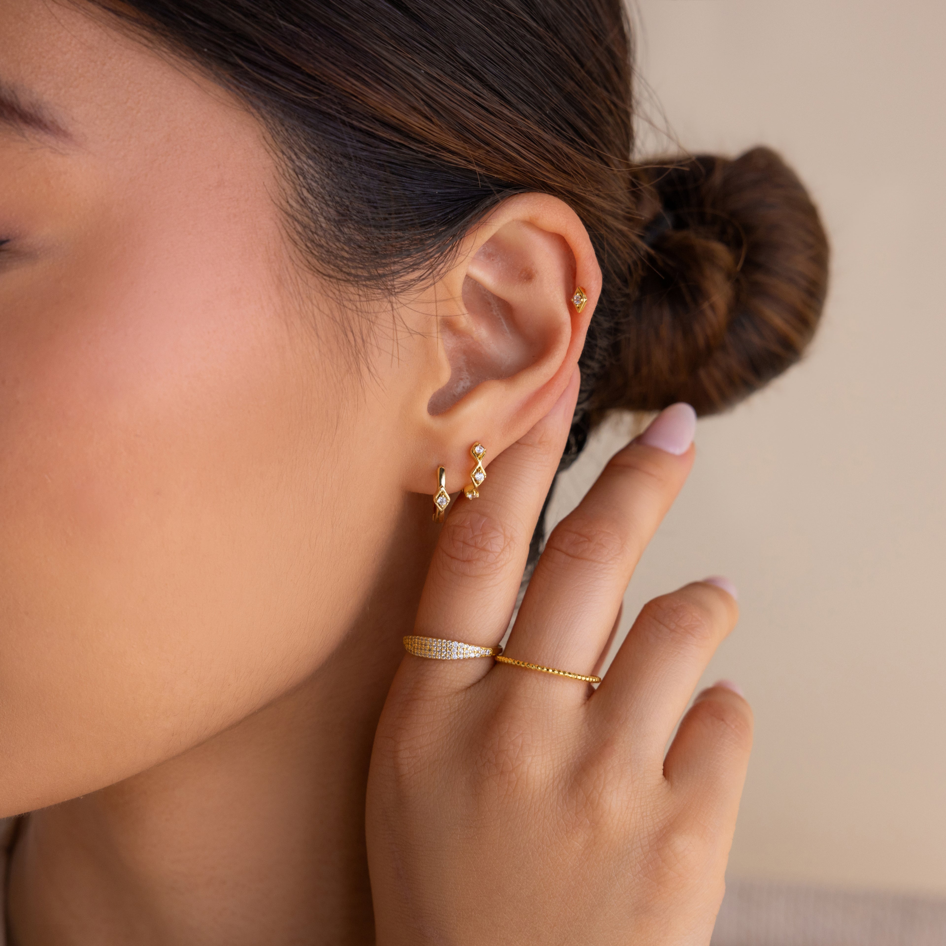 A woman with a neat bun wears the Ericka Diamond Studs, a gold ring, and touches her ear with manicured fingers.