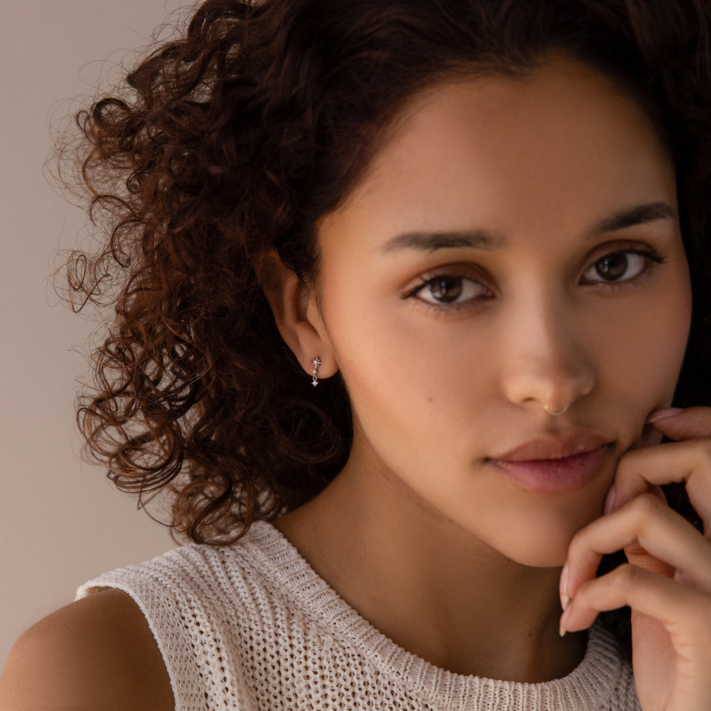 A woman with curly hair in a beige knit top gazes thoughtfully at the camera, hand near her chin, highlighting the elegant Diamond Starburst Drop Studs in Sterling Silver.