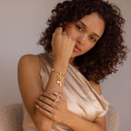 Woman with curly hair poses in a satin top, wearing gold jewelry with pearls and the Hinged Circle Charm Bracelet in 18K Gold, looking at the camera.