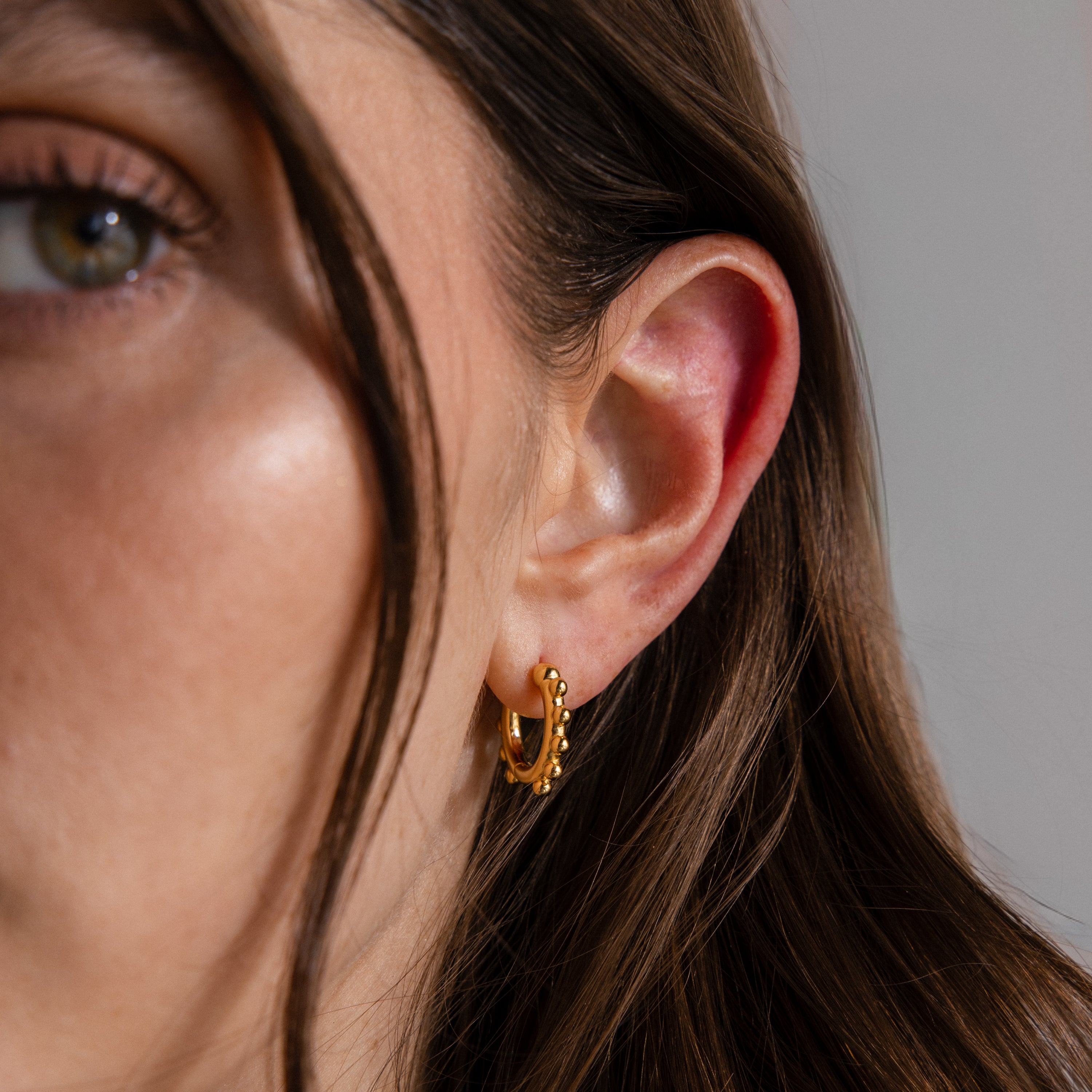Close-up of a woman's ear wearing Beaded Hoops in 18K Gold with textured details, her hair partially covering her face.