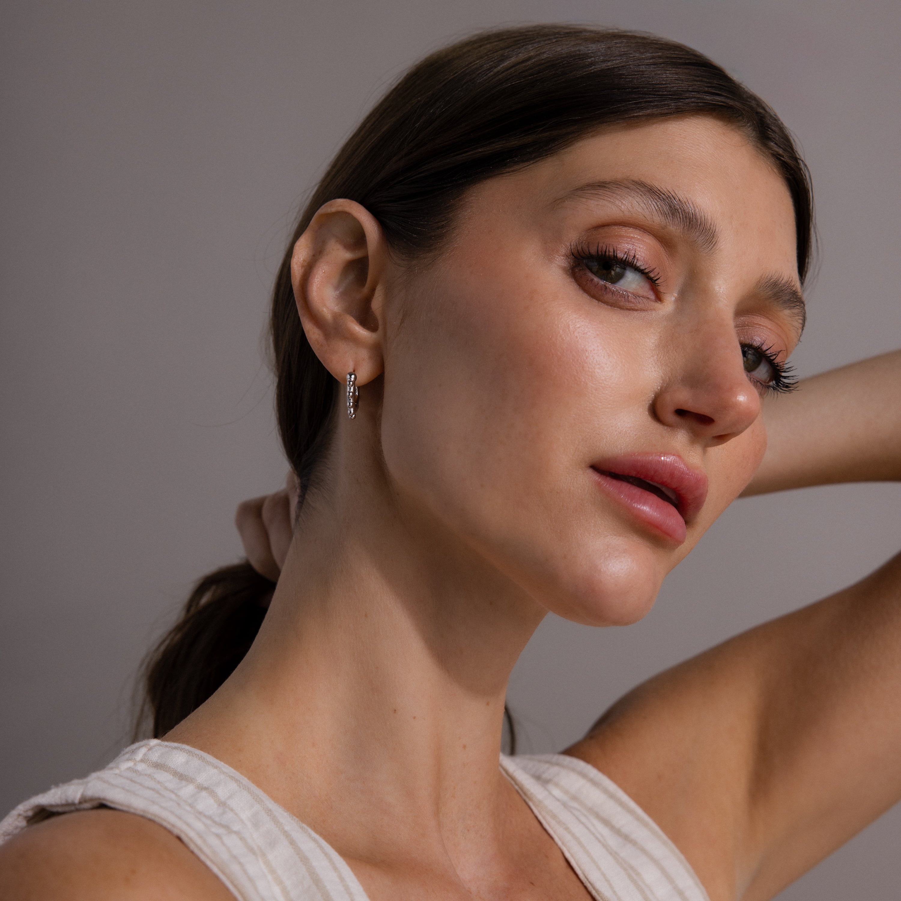 A woman with brown hair in a low ponytail, wearing a sleeveless top and Beaded Hoops in Sterling Silver, poses with her hand behind her head, highlighting a textured glow.