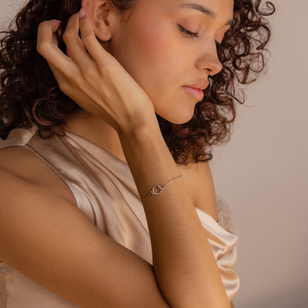 A woman with curly hair touches her neck, highlighting the Hinged Circle Charm Bracelet, which she wears with a light satin top.