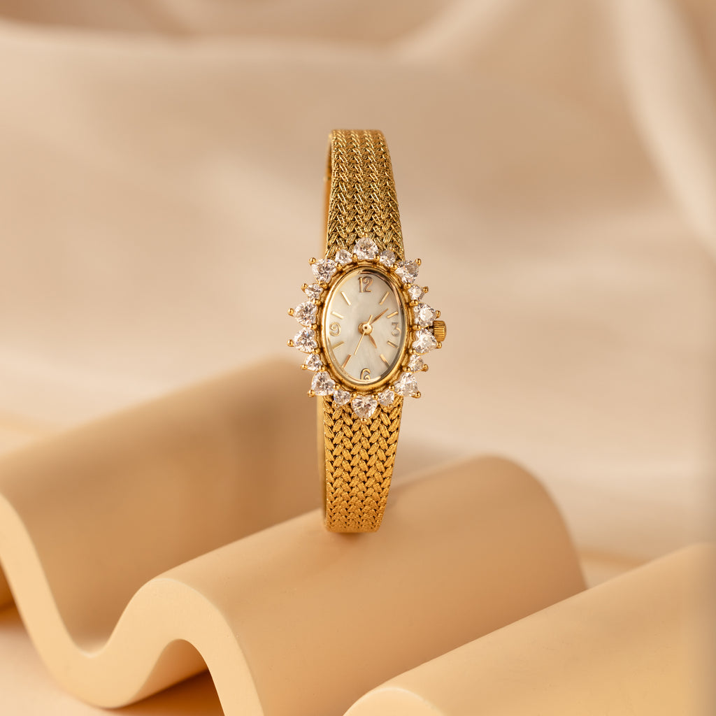 The Pave Heart Timepiece, featuring a jeweled bezel and heart-shaped gemstones, is showcased on a wavy beige surface with a soft background.