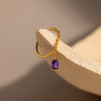 The Amethyst Drop Chain Ring, featuring a delicate gold chain and a small purple gemstone charm, rests on a textured beige surface.