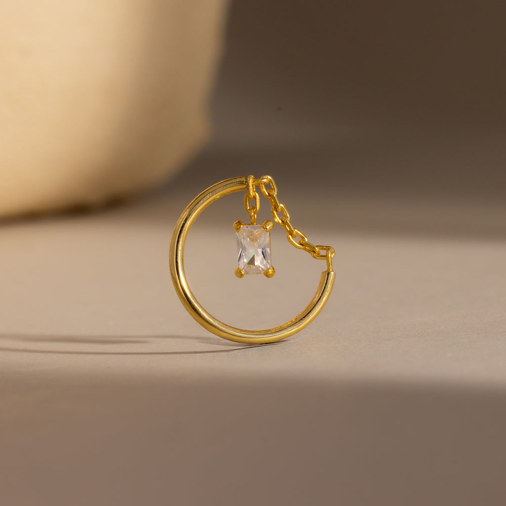 The Birthstone Drop Chain Ring features a gold band with an emerald-cut stone dangling delicately on a chain, showcased against a neutral background.